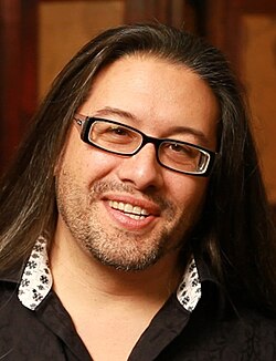 Color photograph of the face of a smiling man with long black hair and glasses
